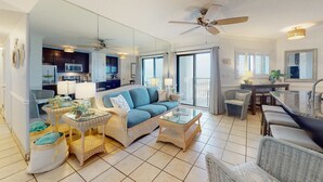 A spacious living area with a sofa, coffee table, and wicker chairs, a kitchen in the background, and a balcony with a view. - Modern condo with Gulf view, shared pool - walk to the beach (Fort Morgan)