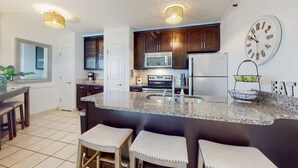 A kitchen with granite countertops, dark wooden cabinets, stainless steel appliances, and a large wall clock. - Modern condo with Gulf view, shared pool - walk to the beach (Fort Morgan)