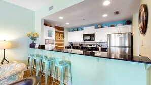 A kitchen with white cabinets, a stainless steel refrigerator, a microwave, and a coffee maker. There are three turquoise bar stools and a vase with yellow flowers on the counter. - Adorable Beach Condo W Shared Pool, Gulf Views, Private Balcony & WiFi (Fort Morgan)