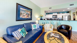 A living room with a blue sofa, a coffee table, and a kitchen area with white cabinets and a stainless steel refrigerator. - Adorable Beach Condo W Shared Pool, Gulf Views, Private Balcony & WiFi (Fort Morgan)