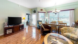 A living room with a flat-screen TV, wooden flooring, a ceiling fan, a dining area with chairs and a table, and a large window with blinds. - Adorable Beach Condo W Shared Pool, Gulf Views, Private Balcony & WiFi (Fort Morgan)