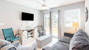 Ein Wohnzimmer mit Flachbildfernseher, Deckenventilator, Sofa, Sessel, kleinem Tisch mit Dekoration und einem Fenster mit Vorhängen.