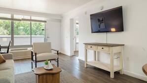 Un salon moderne doté d’un téléviseur à écran plat, d’un fauteuil confortable, d’une petite table ornée d’une plante et d’une grande fenêtre donnant sur un paysage verdoyant.