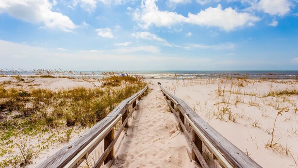 Een houten looppad dat naar een strand met duinen en een heldere lucht leidt.