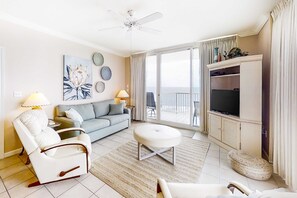 A living room with a sofa, armchair, ottoman, TV, and a balcony with curtains. - Well-appointed condo with amazing views, pool, & hot tub - Close to beach (Fort Morgan)