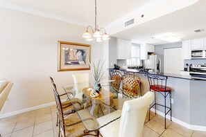 A modern kitchen with white cabinets, a central island, and a dining area with a glass table and wicker chairs. - Beach front condo with amazing views, pool, & hot tub (Fort Morgan)