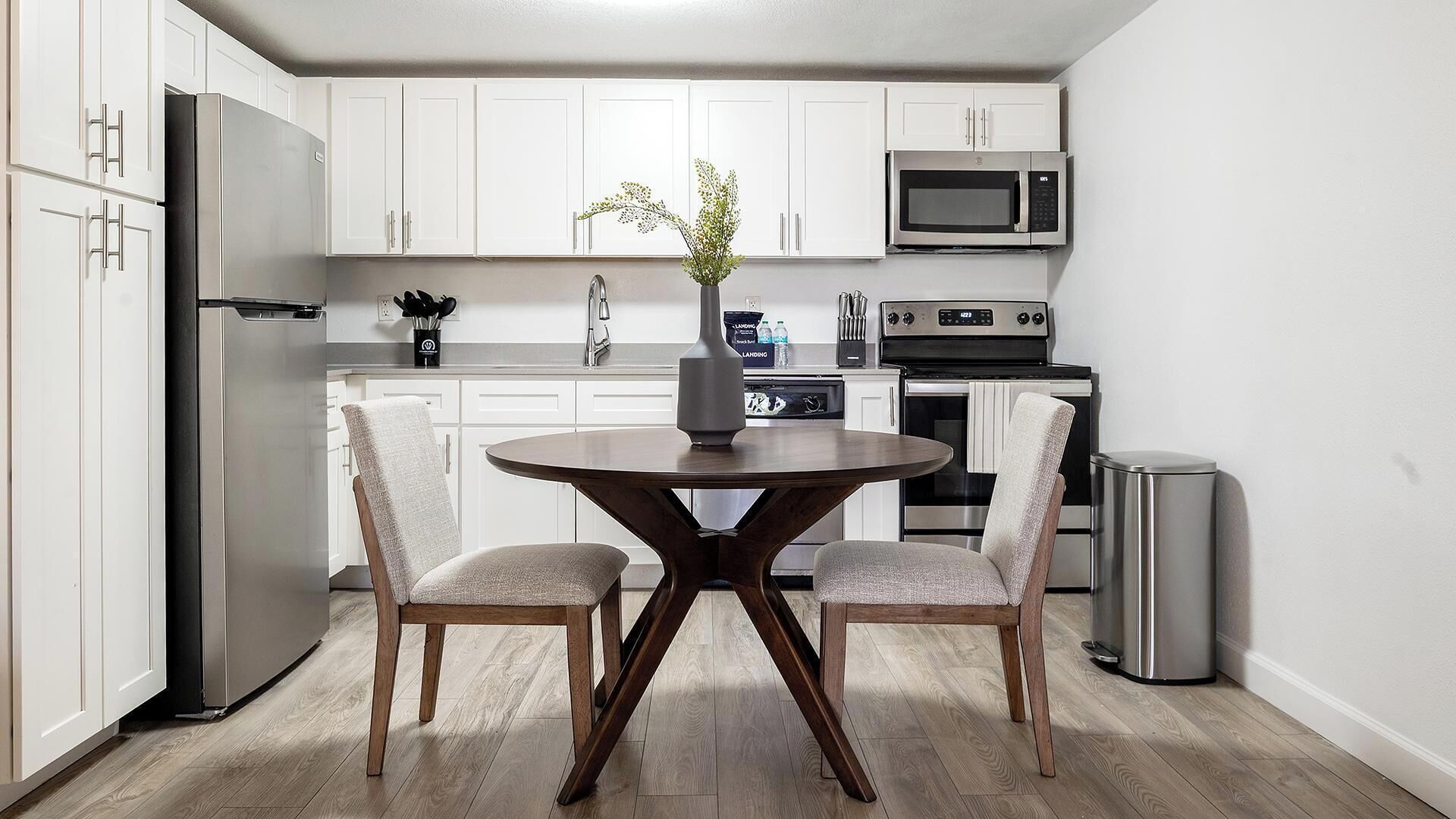 Une cuisine moderne avec des meubles blancs, des appareils en acier inoxydable et un coin repas comprenant une table ronde et deux chaises.