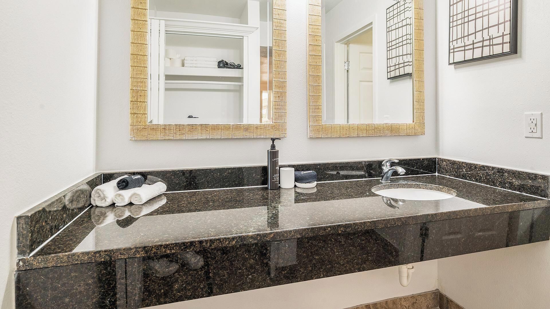 Une salle de bain avec un plan de toilette en granit noir, un lavabo blanc, un miroir avec un cadre doré et un porte-serviettes avec des serviettes.