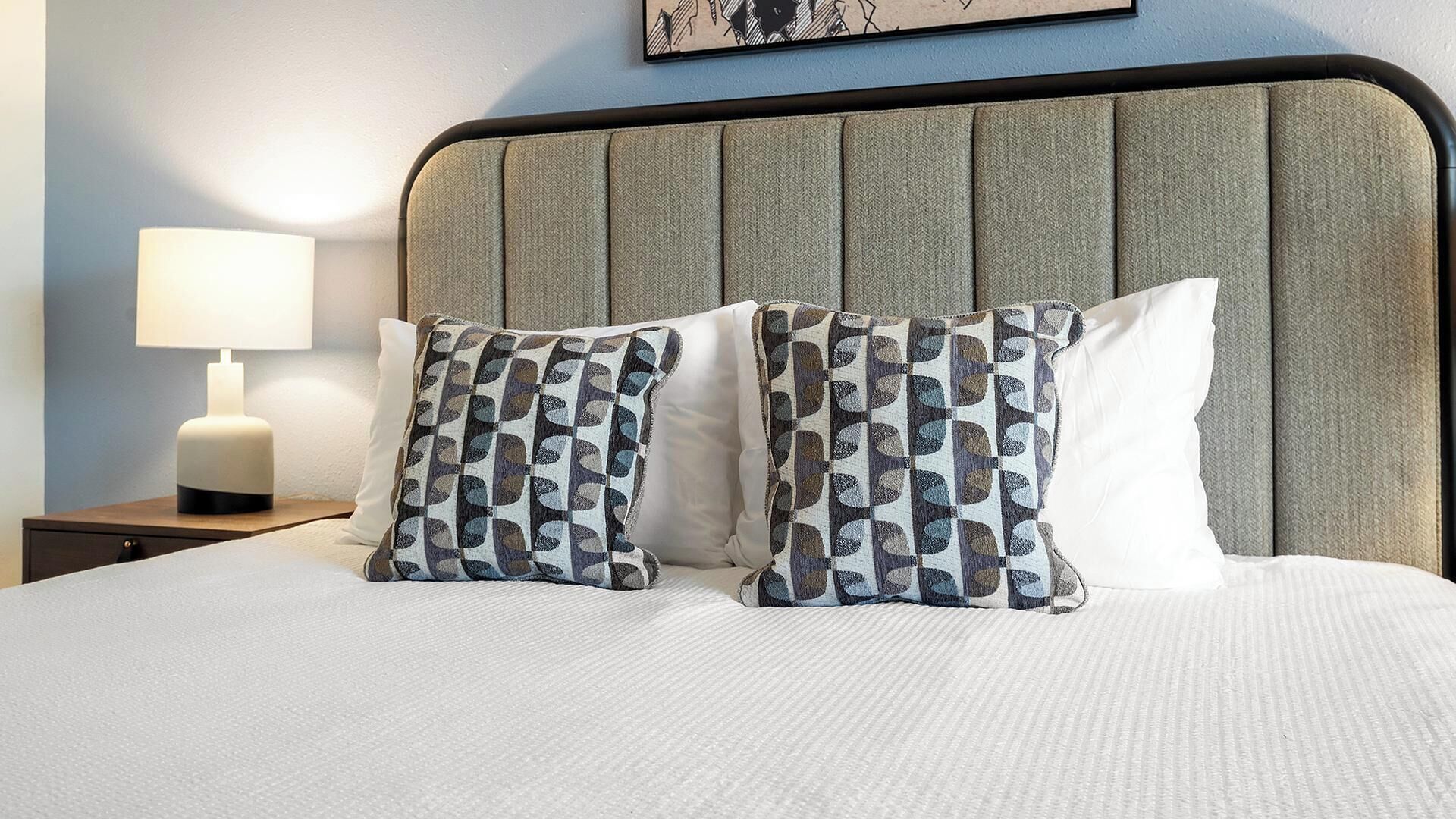 A neatly made bed with patterned pillows, a bedside lamp, and a framed artwork on the wall.