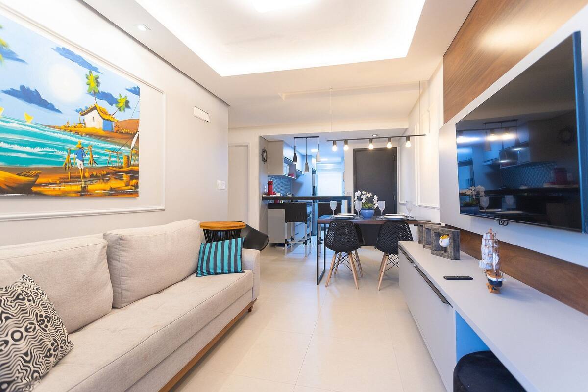 A modern living room with a sofa, a dining area with a table and chairs, a kitchen visible through an open door, and a large painting on the wall.