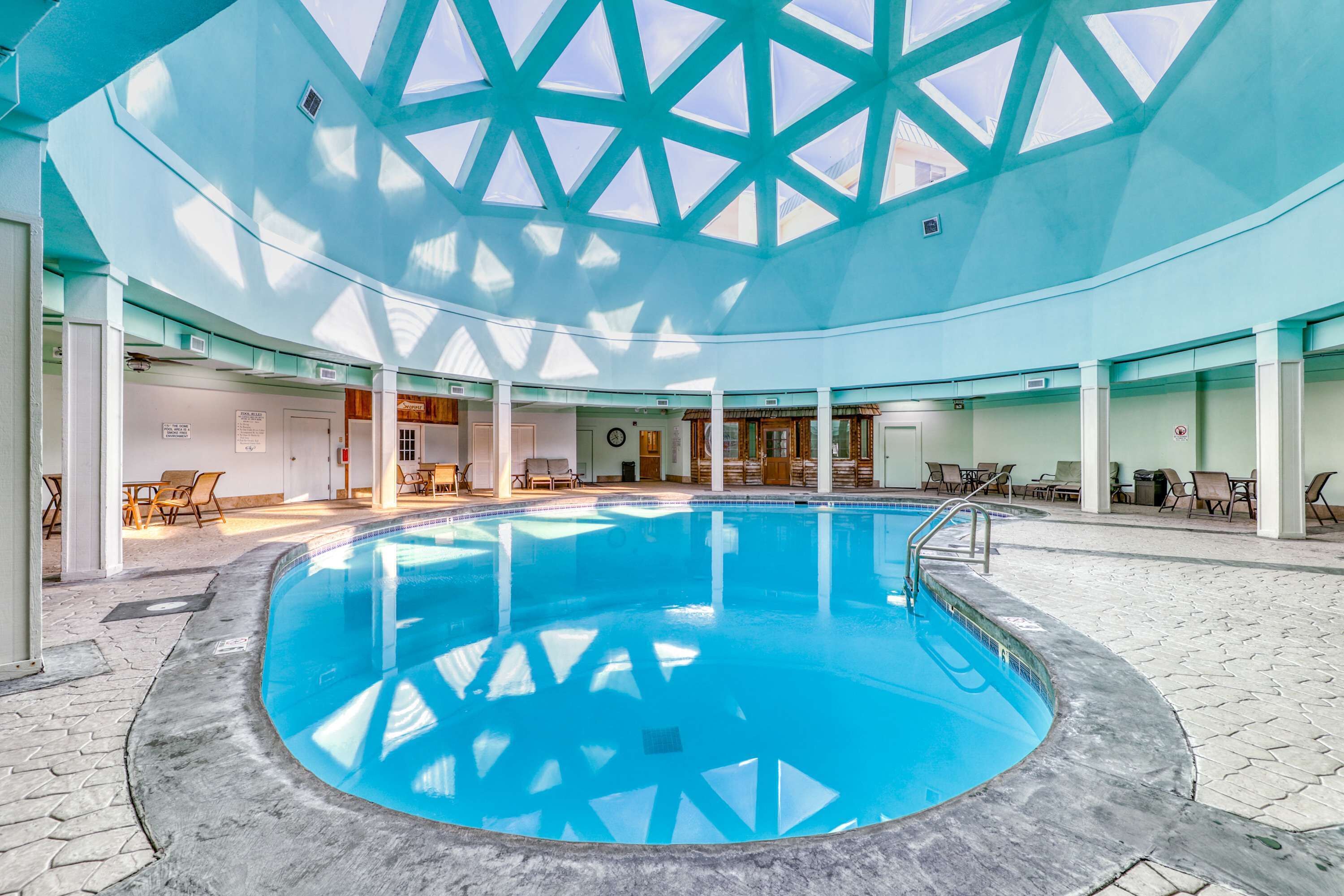 Uma ampla piscina coberta com teto alto e claraboias, cercada por áreas de estar com cadeiras e mesas.