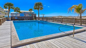 A wooden deck with a swimming pool, palm trees, and a building with a balcony in the background. - Recently Remodeled Condo w/Private Deck, Shared Outdoor Pool, Central AC, WiFi (Fort Morgan)