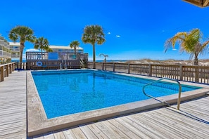 A wooden deck with a swimming pool, palm trees, and a building with a balcony in the background.