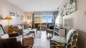 A living room with a sofa, armchairs, a coffee table, and a patterned ottoman. There is a lamp, a television, and artwork on the walls. A sliding glass door leads to an outdoor area with a view of buildings. - Recently Remodeled Condo w/Private Deck, Shared Outdoor Pool, Central AC, WiFi (Fort Morgan)