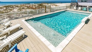 A clear swimming pool with a wooden deck, beach view, and a small building with a porch. - Completely renovated oceanfront home with shared outdoor pool, views, & balcony (Fort Morgan)