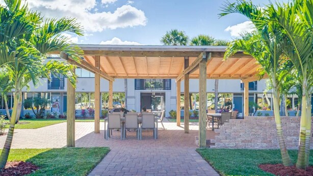 Un patio techado al aire libre con mesas y sillas, rodeado de palmeras y un sendero de ladrillo.