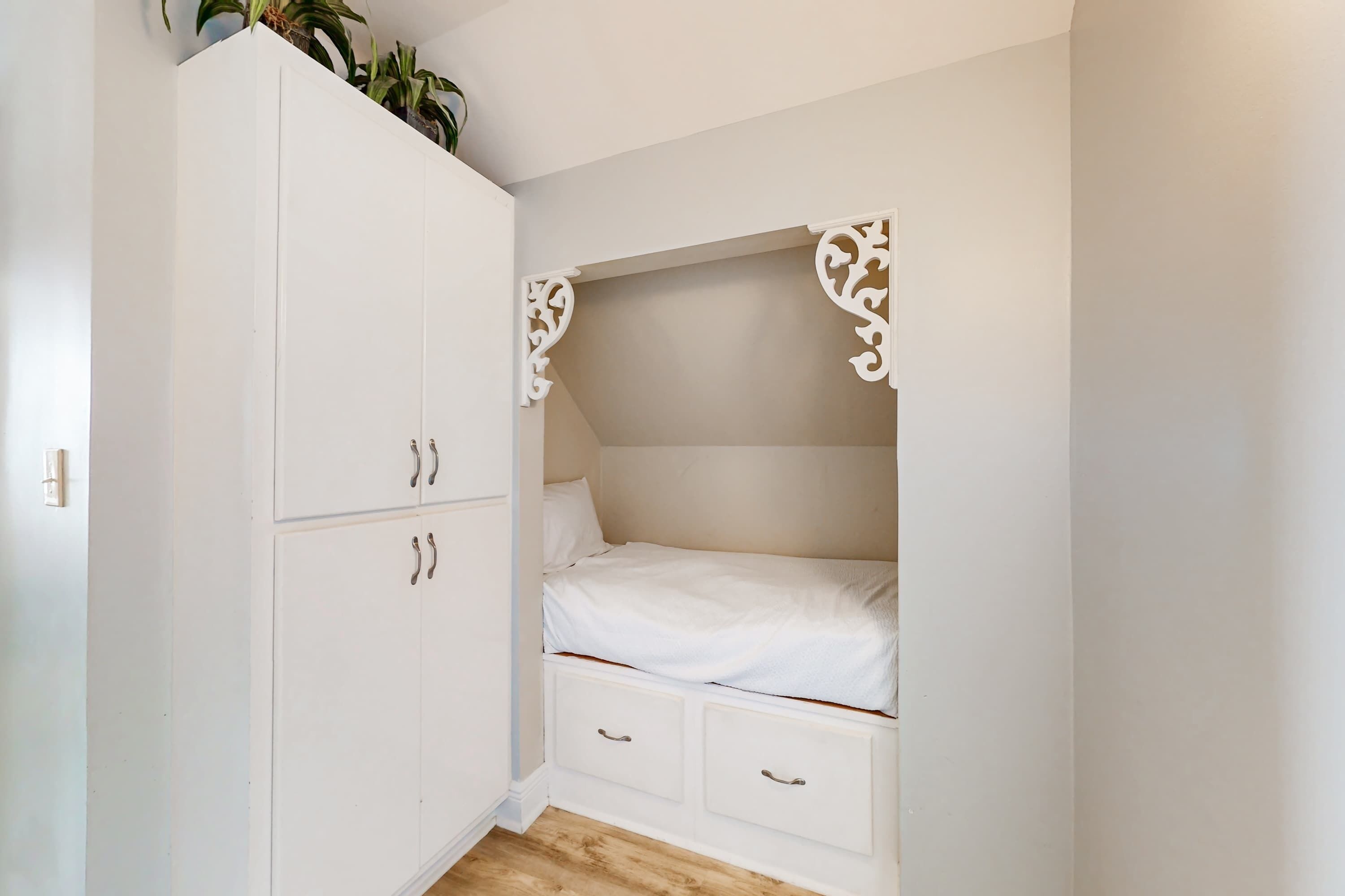 Un dormitorio con un armario blanco, una cama con ropa de cama blanca y una planta pequeña encima del armario.