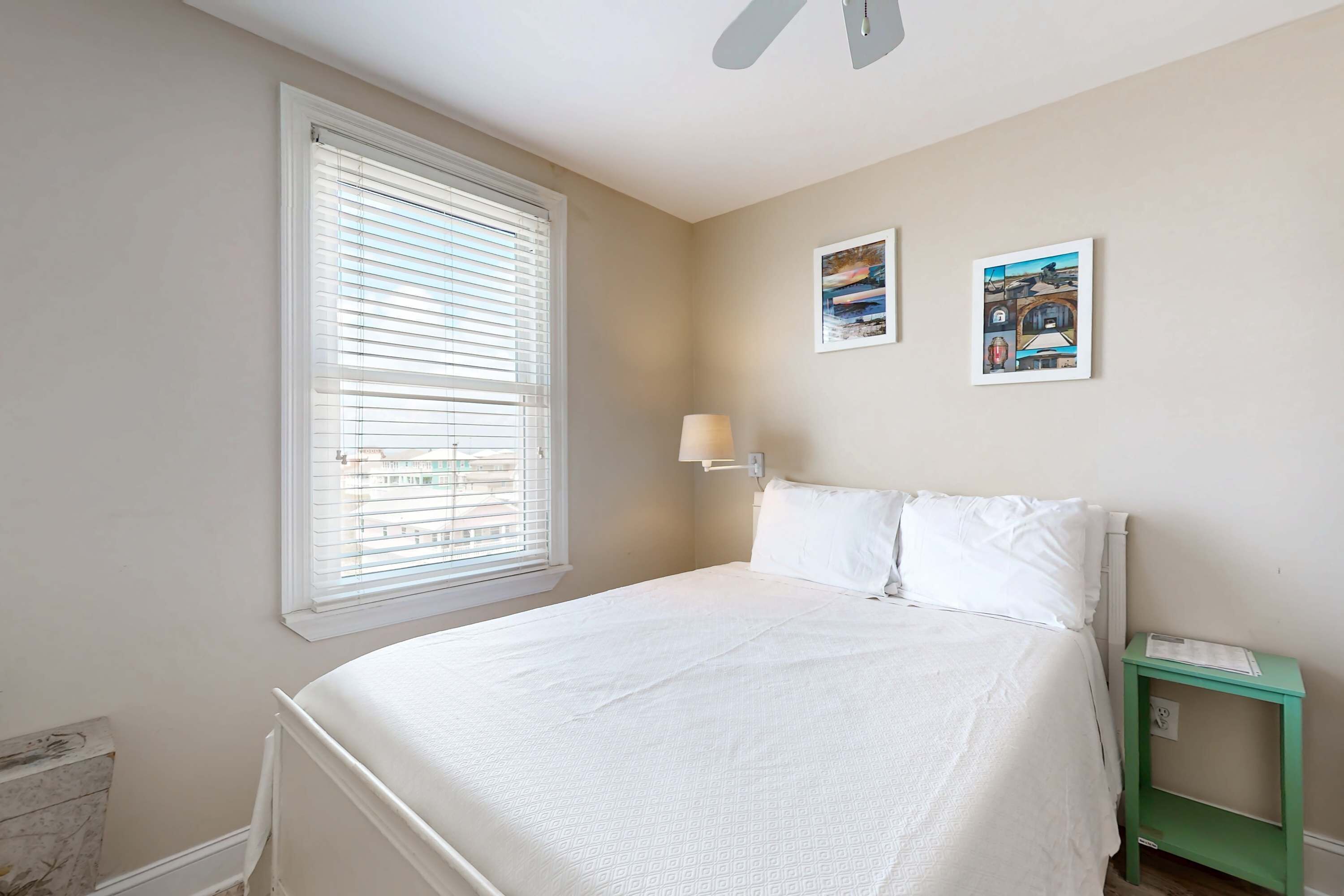 A bedroom with a bed, a window with blinds, a wall-mounted lamp, and two framed pictures.