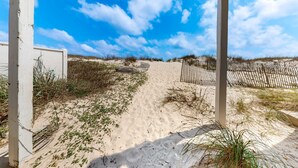 En sti af sand fører til en strand med et træhegn og en klar blå himmel.