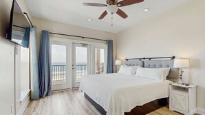 A bedroom with a bed, nightstands, a ceiling fan, a TV, and a balcony with curtains. - Luxury Beachfront Duplex in Fort Morgan | Pool, Elevator and Wedding-Friendly (Fort Morgan)