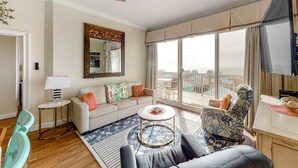 A spacious living room with a large window, a sofa, a coffee table, and a patterned rug. - Ocean-view condo with heated pool, hot tub & rooftop terrace - walk to beach (Fort Morgan)