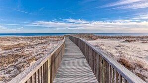 Una pasarela de madera que lleva a la playa, con dunas arenosas y un cielo despejado.