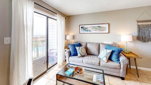 A living room with a sofa, a glass coffee table, a lamp, and a framed picture on the wall. - Renovated Gulf Front Condo with WiFi, Multiple Pools, & Beach Access (Fort Morgan)