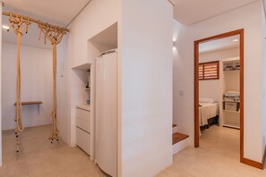 A compact living space with a built-in washer and dryer, a wooden beam with ropes, a white cabinet, and a mirror reflecting a closet with shelves and a bed. - Casa Ladomar em Mundau por Carpediem (Trairi)