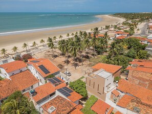 A coastal town with buildings featuring red-tiled roofs, some with solar panels, and a beach with palm trees. - Casa Ladomar em Mundau por Carpediem (Trairi)