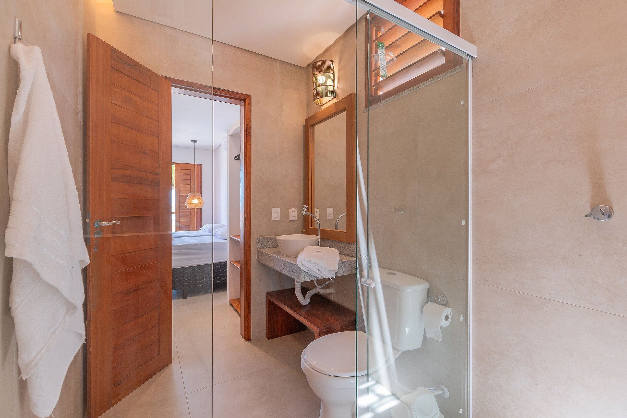 A modern bathroom with a glass shower enclosure, a wooden door, and a visible bedroom in the background.