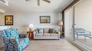 Un salon comprenant un canapé, un fauteuil à motifs, un ventilateur de plafond, un tableau encadré et une vue à travers une grande fenêtre.