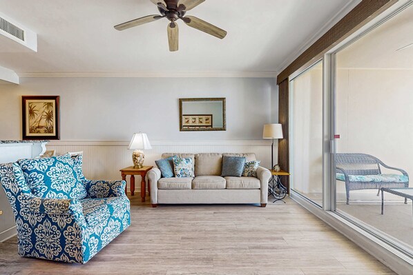 Un salon comprenant un canapé, un fauteuil à motifs, un ventilateur de plafond, un tableau encadré et une vue à travers une grande fenêtre.