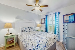 Un dormitorio con cama, mesitas de noche, cómoda, ventilador de techo y ventana con cortinas.