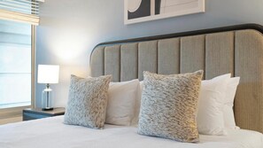 A hotel room with a bed, two pillows, a nightstand, a lamp, and a window with blinds. - Landing | Stunning 1BD, Clubhouse, Gym (Kissimmee)