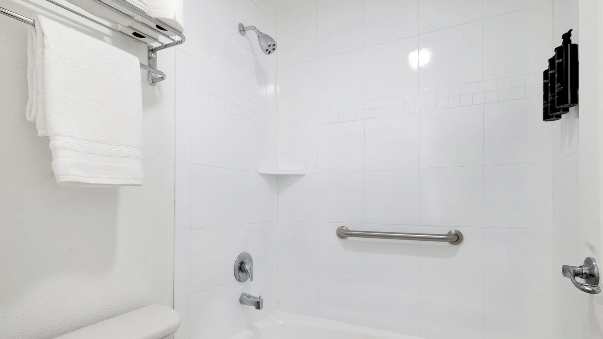 Un baño con pared de azulejos blancos, una ducha con cabezal de ducha manual, un asidero, un inodoro y un perchero para toallas.