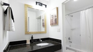 Un bagno con un piano lavabo nero, un lavandino bianco, uno specchio con cornice dorata, una tenda da doccia e un quadro appeso al muro.