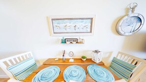 Un comedor con una mesa de madera, sillas blancas, cojines azul listrados, un cuadro enmarcado, un reloj y un letrero que dice 'BISTRO'.