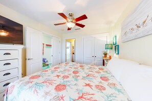 Une chambre à coucher avec un lit, un ventilateur de plafond, des murs blancs et une porte donnant sur une autre pièce.