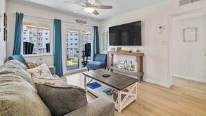 Un salon comprenant un canapé, un fauteuil, une table basse, un téléviseur, un ventilateur de plafond et de grandes fenêtres avec des rideaux.