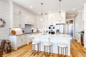 Una cocina moderna con armarios blancos, una isla grande con taburetes, electrodomésticos de acero inoxidable e iluminación colgante.