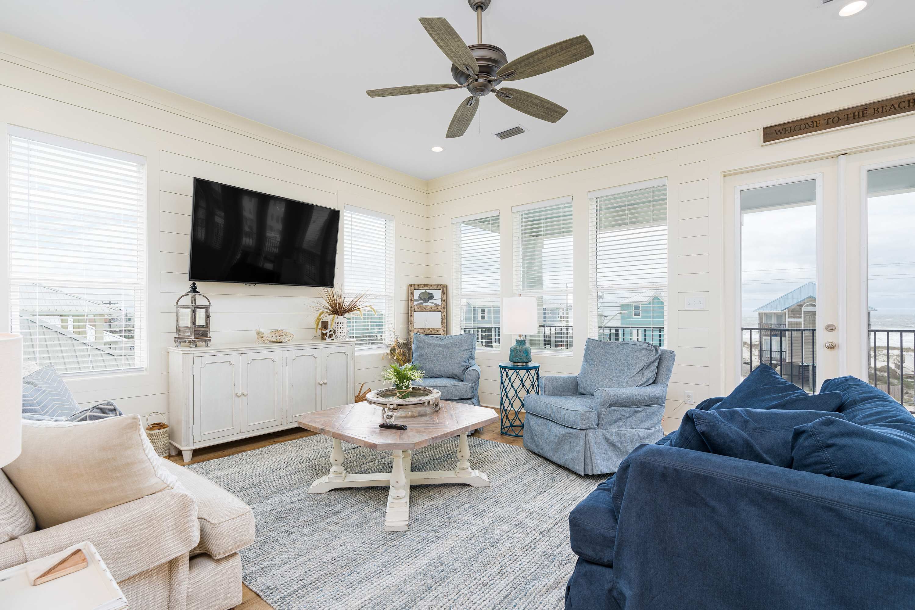 A spacious living room with a ceiling fan, a flat-screen TV, and a view of the city through large windows.