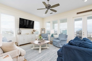 A spacious living room with a ceiling fan, a flat-screen TV, and a view of the city through large windows. - Gorgeous Oceanside Home with WiFi, Private Pool, Central AC, W&D - Steps From Beach (Gulf Shores)