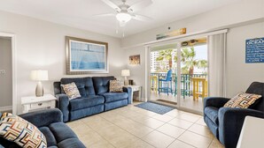 A modern living room with a blue sofa set, a ceiling fan, and a view of a balcony with outdoor furniture. - Oceanwalk 13-305 (New Smyrna Beach)
