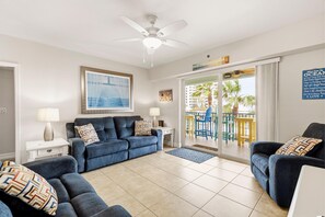 A modern living room with a blue sofa set, a ceiling fan, and a view of a balcony with outdoor furniture. - Oceanwalk 13-305 (New Smyrna Beach)