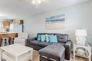 Een woonkamer met een grijze hoekbank, een witte salontafel, een keuken met houten kasten, en een schilderij met een strandthema aan de muur.