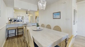 Un espace repas avec une grande table, des chaises, un lustre et une cuisine en arrière-plan.