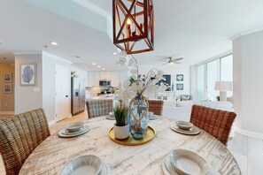 A dining area with a round table, wicker chairs, a hanging light fixture, and a kitchen in the background. - Gulf-front condo with hot tub, sauna, indoor pool & beach access - near dining (Gulf Shores)