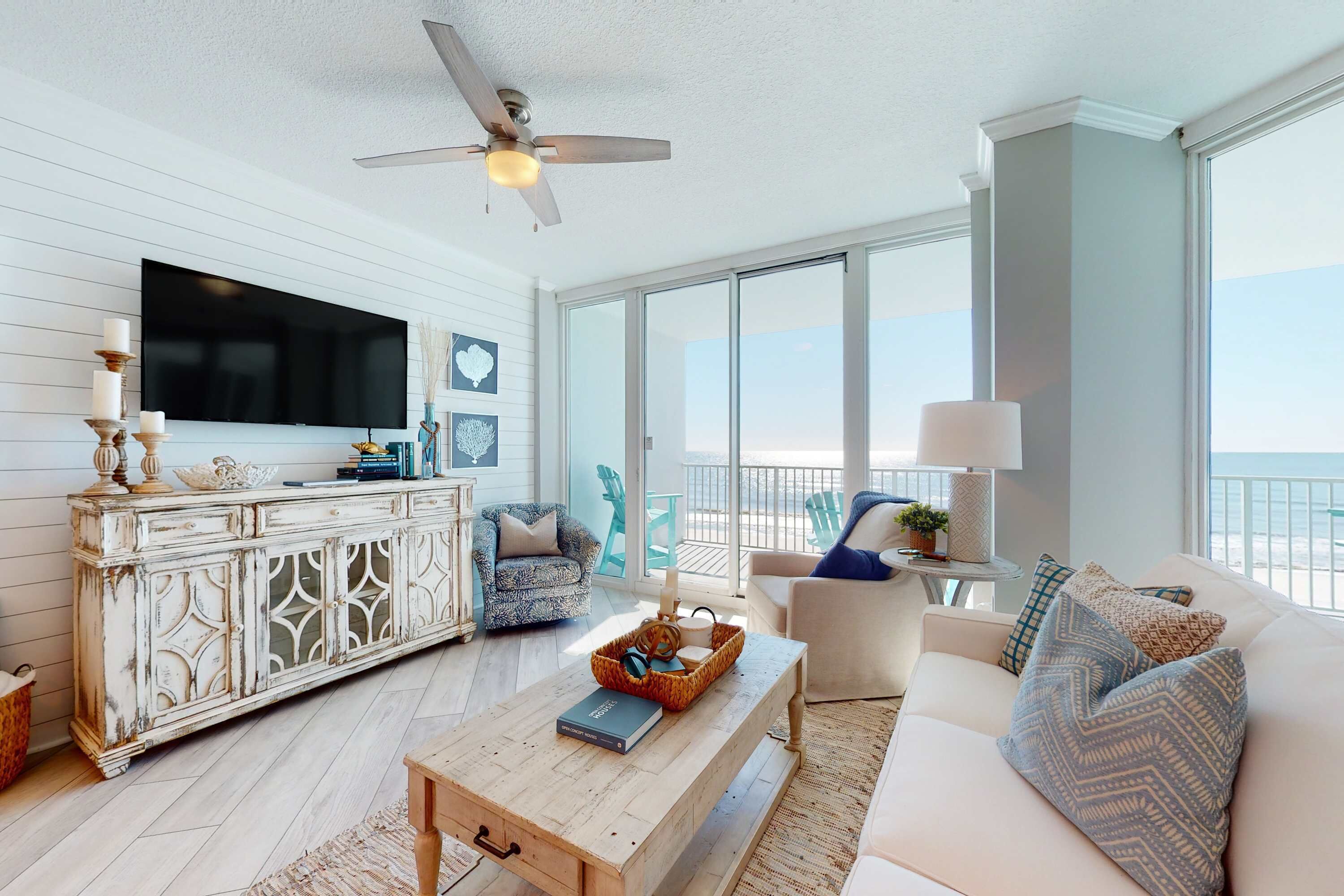 A spacious living room with a large TV, a ceiling fan, a white sofa, a wooden coffee table, and a view of the ocean through large windows.