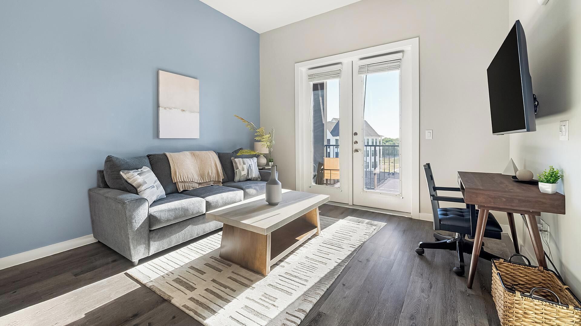 A modern living room with a grey sofa, a coffee table, a TV mounted on the wall, and a balcony visible through the door.