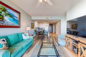 A living room with a teal sofa, a glass coffee table, a dining area with chairs, and a kitchen in the background. - Gulf-front condo w 2 pools, sauna, hot tubs, fitness center & stunning views (Gulf Shores)